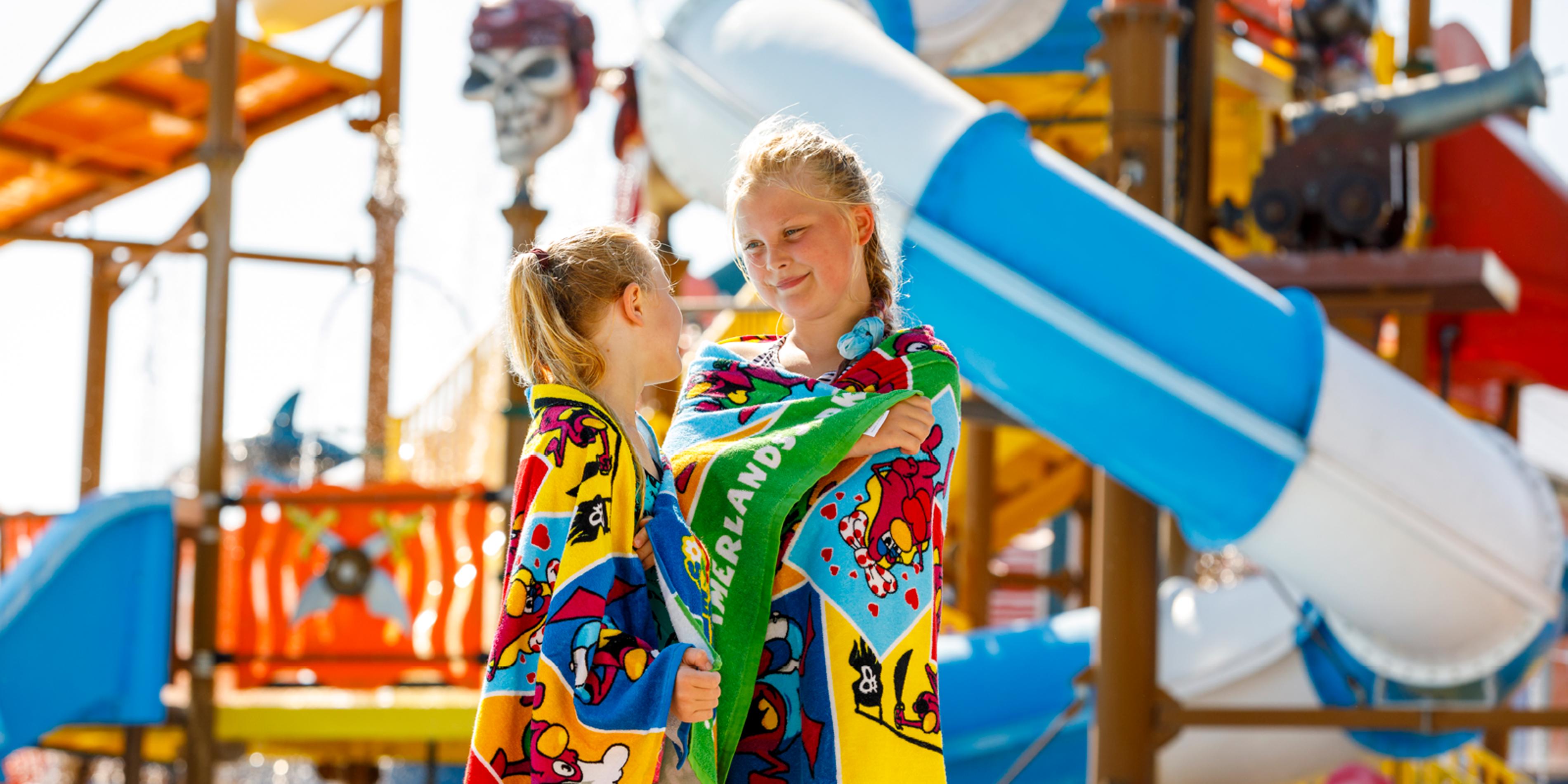 Two girls in colorful bath towels stand in front of a waterslide at a water park.